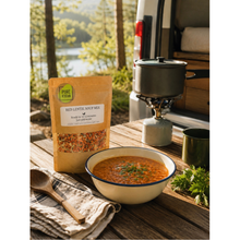 Load image into Gallery viewer, A pouch of Pure Kitten Red Lentil Soup Mix stands on a wooden table beside a bowl of prepared soup, a camping pot on a stove, and an outdoor campsite background with trees and water.
