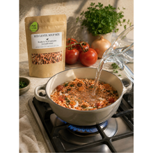 Load image into Gallery viewer, A pouch of Pure Label Red Lentil Soup Mix stands behind a cast-iron pot on a gas stove, with water being poured into the pot containing lentils and vegetables. Tomatoes, a onion, and a potted herb plant are arranged in the background on a kitchen counter.
