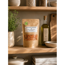 Load image into Gallery viewer, A stand-up pouch of Red Lentil Soup Mix sits on a wooden shelf next to a bottle of olive oil, with kitchen props including a potted herb, bowls, and books in a warm, rustic setting.
