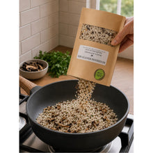Load image into Gallery viewer, A hand holds a kraft paper pouch of Miso Grain Mixture, pouring the loose grains into a black frying pan on a stovetop. The background shows a kitchen setting with a tiled wall and a small bowl of dried mushrooms.

