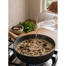 Load image into Gallery viewer, A black non-stick skillet on a gas stove with water being poured into it from a clear glass pitcher, in a kitchen setting with a tiled wall and fresh herbs in the background.
