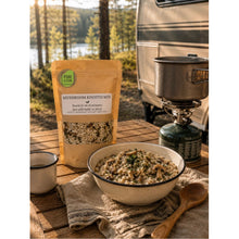Load image into Gallery viewer, A bag of Pure Kitten Mushroom Risotto Mix displayed next to a prepared bowl of risotto on a rustic wooden table, with a camping stove, pot, and a forest backdrop.
